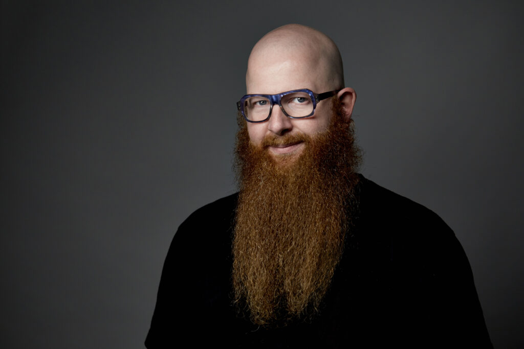 A bald white man with big glasses, a big red beard, and an enigmatic smile